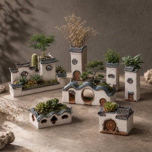 May include: A collection of white ceramic planters shaped like traditional East Asian buildings with blue-grey tiled roofs and brown accents. The planters feature various architectural details including arched doorways, circular windows, and tiered roof sections.