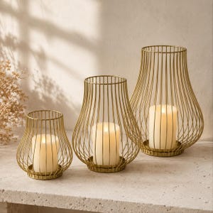 May include: Three gold-colored metal wire candle holders of varying heights, each featuring a rounded, tapered cage design with vertical bars and a circular base. Inside each holder sits a single white pillar candle with a warm, glowing light.