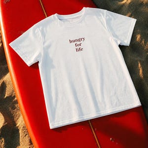 Peut inclure: Un t-shirt blanc à manches courtes avec le texte "hungry for life" imprimé en police serif rouge foncé au centre de la poitrine. Le vêtement présente une encolure ronde et une coupe standard, offrant un design épuré sur un tissu blanc.