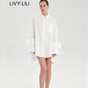 May include: A white button-down shirt with a classic collar, a single chest pocket, and long sleeves featuring wide, flared, structured cuffs. The shirt has a high-low hemline and the text LIVY LILI is printed in black on the upper left side.
