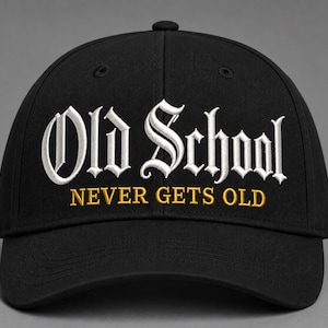 May include: A black baseball cap featuring white embroidered text that reads Old School in a gothic font, with the words NEVER GETS OLD embroidered in gold underneath. The cap has a structured crown, a curved brim, and a top button.