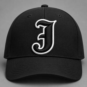 May include: A black baseball cap featuring a prominent, embroidered letter J in a stylized gothic font. The letter is black with a thick white border, centered on the front panel of the cap. The cap has a structured crown and a curved brim.