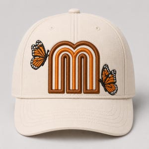 Könnte beinhalten: Eine cremefarbene Baseballkappe mit einem gestickten Motiv auf der Vorderseite. Das Design zeigt einen großen, stilisierten Buchstaben M in Braun- und Orangetönen, flankiert von zwei Monarchfaltern mit orangefarbenen, schwarzen und weißen Flügeln.