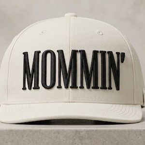 May include: A cream-colored baseball cap featuring the word MOMMIN' embroidered in bold, black, raised lettering across the front panels. The cap has a structured crown, a flat brim, and a small fabric tag visible on the side panel.