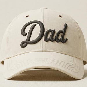 Op de afbeelding: Een crèmekleurige baseballpet met het woord Dad geborduurd in donkerbruine, verhoogde schrijfletters op de voorpanelen. De pet is voorzien van een gebogen klep, stikseldetails en metalen oogjes op de kroon voor extra ventilatie.