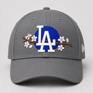 Puede incluir: Una gorra de béisbol gris con un círculo azul bordado y las letras blancas LA en el centro. Una rama marrón con flores de cerezo blancas y rosas se extiende horizontalmente sobre el logotipo. Un pequeño logotipo de bandera blanca está en el lateral.