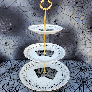 May include: A three-tiered serving stand featuring white ceramic plates with black celestial and tarot card illustrations. The plates are connected by a central gold-colored metal rod with a decorative loop handle at the very top of the structure.
