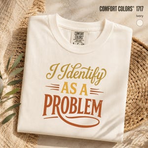 May include: An ivory-coloured crew neck t-shirt featuring the text "I Identify AS A PROBLEM" printed in a gradient of gold, yellow, and burnt orange. The shirt includes a Comfort Colors brand label on the inside collar, indicating size S, P, CH.