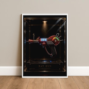 May include: A framed print featuring a red and metallic ray gun displayed inside a dark glass case. The ray gun has glowing blue lights and a circular gauge with green, yellow, and red segments. The text RAY GUN is printed on a plaque below.