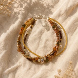 Pode incluir: Uma pulseira de várias camadas com contas em tons de âmbar, castanho, creme e translúcidas de várias formas e tamanhos. A pulseira inclui uma conta em forma de gota e um fecho de mosquetão em metal prateado com uma corrente extensora.