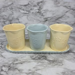 May include: A set of three ceramic cups arranged on a matching rectangular tray. Two cups are pale yellow and one central cup is light blue. Each cup features a subtle embossed floral vine pattern and a wavy rim. The tray has a matching wavy edge.
