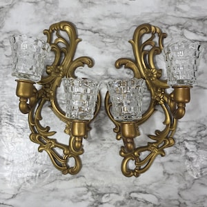 May include: A pair of wall-mounted candle holders featuring ornate gold-toned scrollwork frames with floral accents. Each holder supports two clear, textured glass votive cups with a grid-like pattern, designed to hold candles securely.