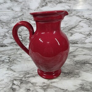May include: A glossy, deep red ceramic pitcher featuring a rounded body, a wide flared rim, and a curved handle on the side. The pitcher has a smooth, reflective surface and sits on a circular pedestal base, showcasing a solid, vibrant crimson color.
