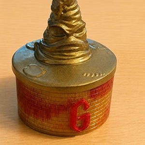 May include: A round container with a brick-patterned base in shades of orange and red, featuring a prominent red letter G on the front. The lid is metallic gold and topped with a sculpted, pointed hat figure with deep folds and a facial expression.
