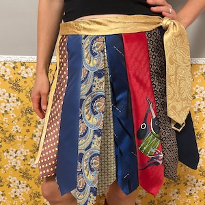 May include: A skirt constructed from multiple neckties in various patterns and colours, including red, blue, gold, and black. The skirt features a wide, gold-coloured paisley waistband that ties at the side, with the individual neckties hanging vertically.