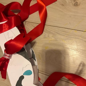 Może przedstawiać: Biała wycinanka z papieru w kształcie maski z ręcznie rysowanymi czarnymi i niebieskimi kształtami, ozdobiona długimi, jaskrawoczerwonymi satynowymi wstążkami zawiązanymi wokół konstrukcji. Maska posiada małe biało-czerwone zdobienie przy węźle.