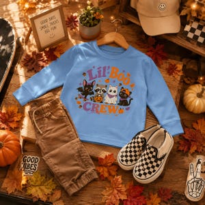 Könnte beinhalten: Ein hellblaues Langarmshirt mit einer Grafik aus Fledermaus, Waschbär, Geist und Katze sowie dem Text Lil Boo Crew, kombiniert mit einer braunen Hose und schwarz-weiß karierten Slip-on-Schuhen. Die Grafik zeigt Kürbisse und Herbstblätter.
