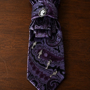 Può includere: Una cravatta con motivo paisley viola e nero caratterizzata da una spilla a cammeo color argento sul nodo e quattro piccoli ciondoli a forma di chiave antica color argento fissati sulla parte anteriore. La cravatta ha un'estremità a punta e un design pieghettato.