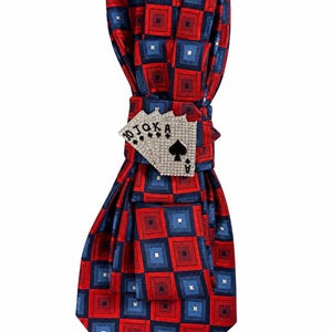 May include: A necktie featuring a repeating geometric pattern of red and blue squares with a central silver-toned metal accessory shaped like a fan of four playing cards, including a 10, Jack, Queen, King, and an Ace of Spades, all encrusted with clear crystals.