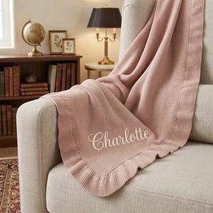 May include: A dusty rose knitted blanket with a ruffled border and the name Charlotte embroidered in white script lettering. The blanket is draped over a light-colored upholstered armchair, featuring a textured knit pattern throughout the fabric.