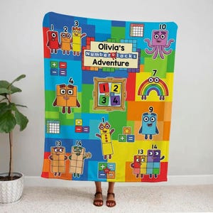 Peut inclure: Une couverture rectangulaire en polaire présentant un motif à grille coloré avec des personnages Numberblocks, les chiffres de 1 à 14, des symboles mathématiques et un cadre central avec le texte Olivia's Numberblocks Adventure en noir.