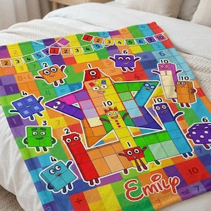 Peut inclure: Une couverture en polaire colorée présentant un motif quadrillé avec des chiffres et des symboles mathématiques. Le design inclut divers personnages en forme de blocs avec des visages, une grande étoile centrale et le prénom Emily.