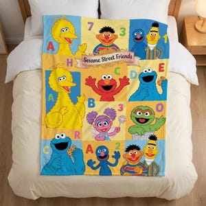 Op de afbeelding: Een rechthoekige plaid met een raster van Sesame Street-personages, waaronder Big Bird, Elmo, Cookie Monster, Abby Cadabby, Bert, Ernie en Grover. Het ontwerp bevat letters, cijfers en de tekst Sesame Street Friends.