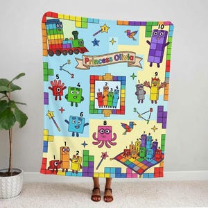 Peut inclure: Une couverture rectangulaire en polaire avec un fond bleu clair et jaune, illustrée de personnages Numberblocks colorés, d'un train et de formes géométriques. Une bannière centrale porte le texte Princess Olivia entouré de chiffres.