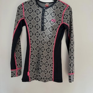 May include: A long-sleeve henley shirt featuring a grey and black snowflake pattern on the front and sleeves, accented by solid black side panels and bright pink piping. The shirt has a four-button placket and a small pink logo on the chest.