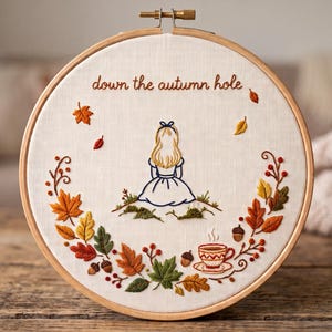 Op de afbeelding: Een ronde houten borduurring met witte stof en een handgeborduurd ontwerp. De tekst "down the autumn hole" is geborduurd boven een figuur met blond haar. Een krans van kleurrijke herfstbladeren, eikels en een theekopje omringt het geheel.