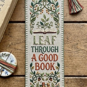 May include: A rectangular fabric bookmark with beige linen, featuring hand-embroidered green vines, orange berries, and a stack of books. The text reads LEAF THROUGH A GOOD BOOK in green and orange thread. It has a green braided cord and tassel.