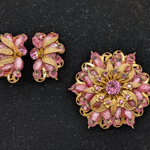 May include: A gold-tone jewelry set consisting of a large circular brooch and a pair of matching earrings. Each piece features textured gold-tone metal leaves interspersed with translucent pink glass beads and small pink faceted rhinestones.
