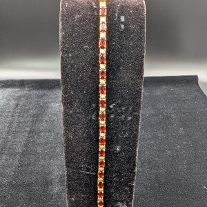 May include: A gold-tone tennis bracelet featuring a series of oval-cut deep red garnet gemstones alternating with small clear white stones. The bracelet is constructed with a flexible metal link chain and a secure clasp mechanism at the bottom end.
