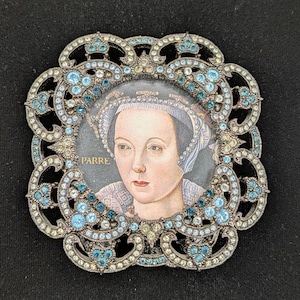 May include: A square brooch with rounded, ornate edges featuring a portrait of a person wearing a headpiece and the text PARRE. The frame is decorated with numerous small, light blue and clear crystals set in a dark, metallic, filigree-style metal.