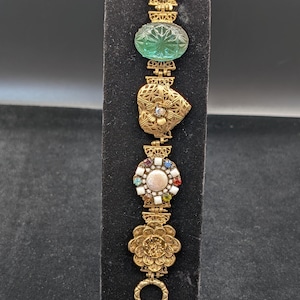 May include: A gold-tone link bracelet featuring a series of decorative charms including a floral top piece, a translucent green oval glass cabochon, a filigree heart, a circular medallion with multi-colored stones, and a detailed flower charm.