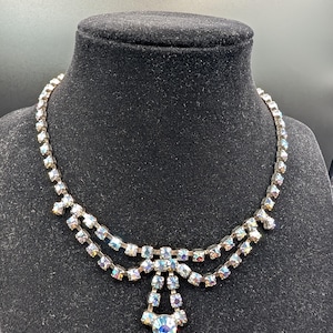 May include: A double-strand necklace featuring iridescent, multi-colored rhinestones set in silver-toned metal. The design includes a central drop element with a larger circular rhinestone at the bottom, connected by smaller stones in a tiered arrangement.