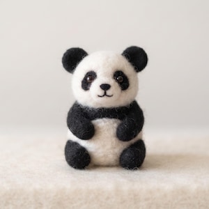 Peut inclure: Une petite figurine de panda en laine feutrée à l'aiguille, présentant un corps blanc avec des oreilles, des taches oculaires, des membres et un nez noirs. Le panda a des yeux ronds sombres et une bouche cousue, en fibres de laine texturées.
