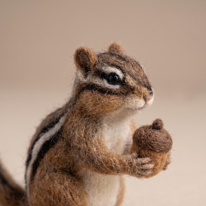 May include: A needle-felted chipmunk figurine crafted from brown, tan, and white wool fibers. The small animal features distinct dark brown and white stripes along its back and head, with black eyes and a small brown acorn held in its front paws.