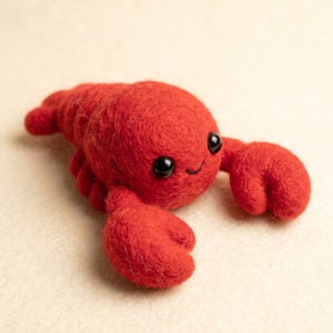 Peut inclure: Une petite figurine de homard en laine feutrée rouge vif avec un corps arrondi, deux pinces avant proéminentes et deux petits yeux en perles noires brillantes. La texture du homard est duveteuse et douce, avec un sourire cousu sous les yeux.