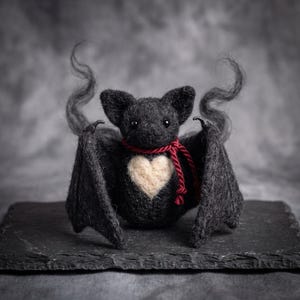 Può includere: Una statuetta di pipistrello nera in lana infeltrita con finitura testurizzata, caratterizzata da una toppa a forma di cuore color crema sul petto e un sottile cordino rosso annodato al collo. Presenta orecchie a punta, ali spiegate e antenne.