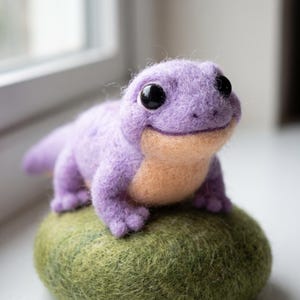 Könnte beinhalten: Eine nadelgefilzte violette Echsenfigur mit einem hellpfirsichfarbenen Bauch und großen schwarzen Augen. Die Echse steht auf einer strukturierten, runden, moosgrünen Filzbasis. Das Wesen hat einen glatten, abgerundeten Kopf und kleine Füße.