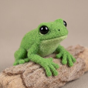 May include: A needle-felted green frog figurine with a textured wool surface, large glossy black eyes, and four distinct toes on each foot. The frog is positioned in a resting pose, perched atop a cylindrical piece of light brown textured wood.
