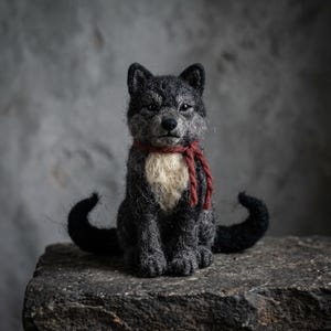 May include: A needle-felted wolf figurine crafted from dark gray and black wool fibers. The wolf features a cream-colored chest patch, pointed ears, and a dark nose. A thin, braided dark red scarf is tied around its neck. The wolf sits upright.