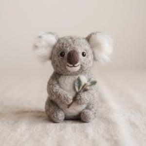 Könnte beinhalten: Eine nadelgefilzte Koala-Figur aus grauer und weißer Wolle. Der Koala hat große, flauschige weiße Ohren, eine dunkelbraune Nase und schwarze Augen. Er sitzt in einer aufrechten Position und hält einen kleinen grünen Zweig mit Eukalyptusblättern.