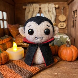 May include: A needle-felted vampire figurine with a pale face, black hair, and large black eyes. It wears a black cape with a red lining and features two small white fangs. The figure is positioned in a miniature room setting with pumpkins.