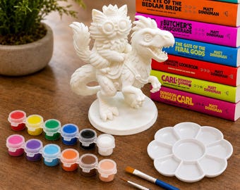 May include: A white unpainted figurine of a cat wearing goggles and a crown riding a dinosaur, accompanied by a strip of twelve small paint pots, two paintbrushes, and a white flower-shaped plastic palette, next to a stack of six colorful books.
