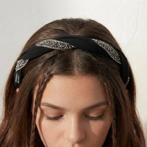 May include: A black fabric headband featuring a twisted design with two integrated bands of shimmering silver-toned crystals. The accessory has a smooth matte finish on the fabric sections and includes a small black label with white text.