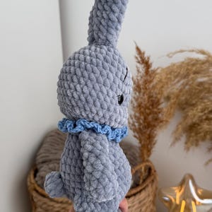 May include: A gray crocheted plush rabbit toy made from soft, chunky yarn. The rabbit features long, upright ears, a small round tail, and a ruffled light blue collar around its neck. It has a smooth, textured surface and a dark, round eye.