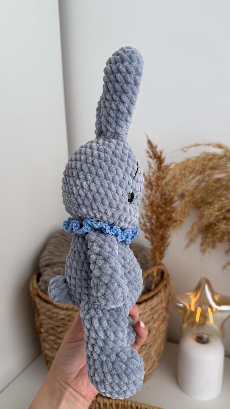 May include: A gray crocheted plush rabbit toy made from soft, chunky yarn. The rabbit features long, upright ears, a small round tail, and a ruffled light blue collar around its neck. It has a smooth, textured surface and a dark, round eye.