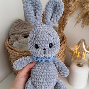 May include: A handmade crochet plush toy in the shape of a rabbit, crafted from soft grey yarn. The rabbit features long upright ears, black plastic eyes, a small stitched nose, and a light blue ruffled collar around its neck.